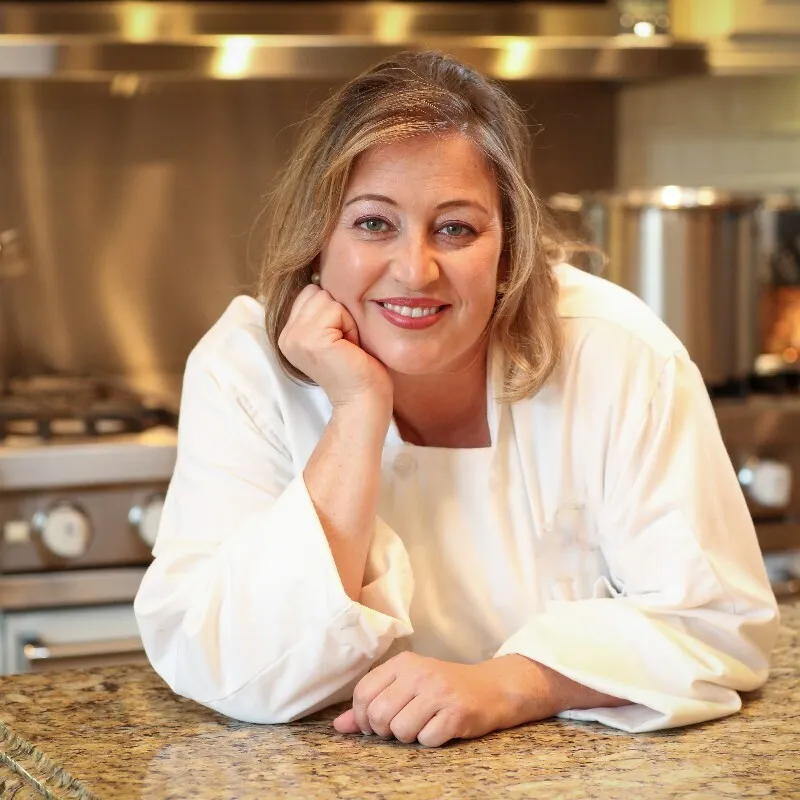 A woman with light brown hair smiling in a white chef's coat, leaning on a granite countertop in a commercial kitchen.
