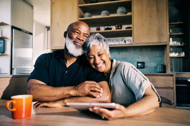 An older couple sits closely together at a kitchen table, smiling and holding a tablet, with a coffee mug in front of them.