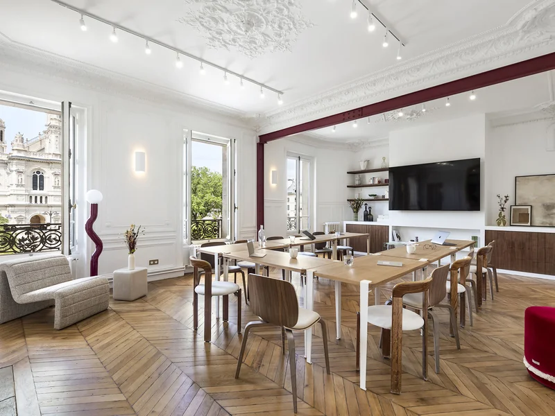 Salon lumineux et minimaliste avec sol en bois clair, table carrée en bois entourée de quatre chaises, fauteuil beige, canapé clair avec table basse blanche, éléments décoratifs noirs, grande fenêtre laissant entrer la lumière naturelle et table avec nappes blanches présentant des pâtisseries, fruits et cafetière.