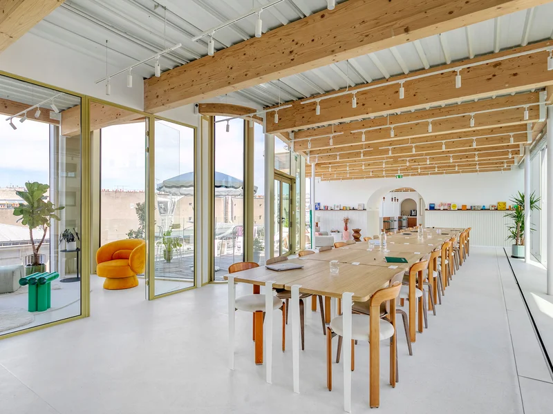 Espace de réunion lumineux avec une longue table en bois entourée de chaises en bois clair, un plafond avec poutres apparentes en bois, de grandes fenêtres donnant sur une terrasse avec une chaise orange et des plantes.