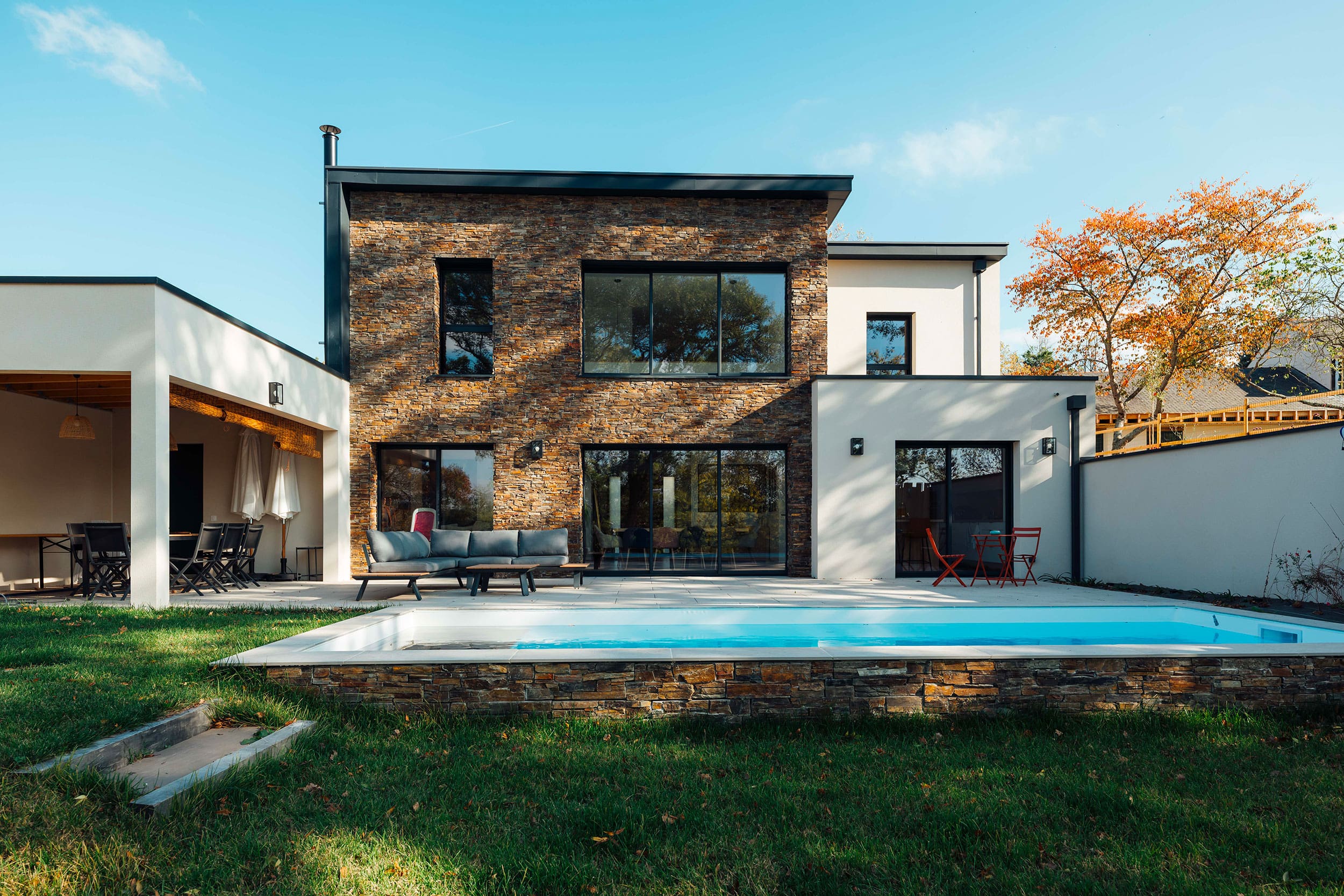 Maison moderne avec façade en pierre et piscine dans le jardin avec mobilier extérieur sous un ciel bleu.