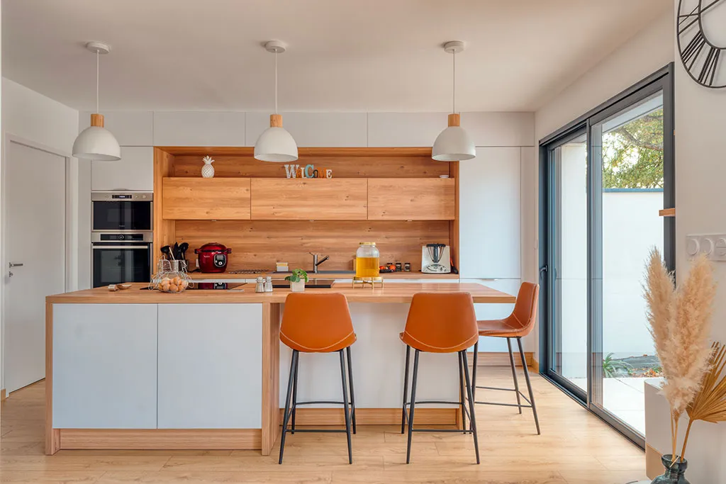 Cuisine moderne avec îlot en bois clair, trois tabourets orange, électroménagers intégrés et grandes baies vitrées laissant entrer la lumière naturelle.