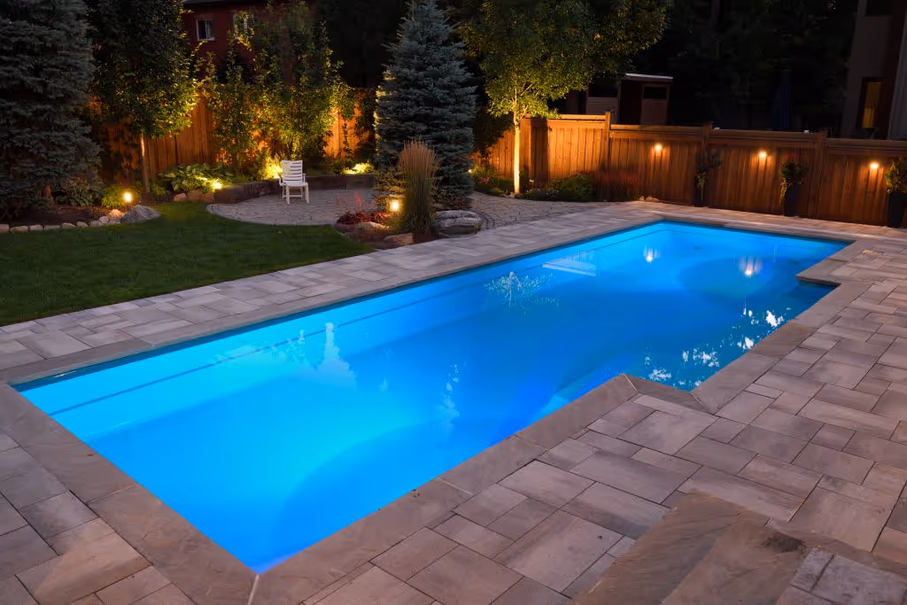 A backyard swimming pool at night. Lights illuminate the entire space, including the pool and fence.