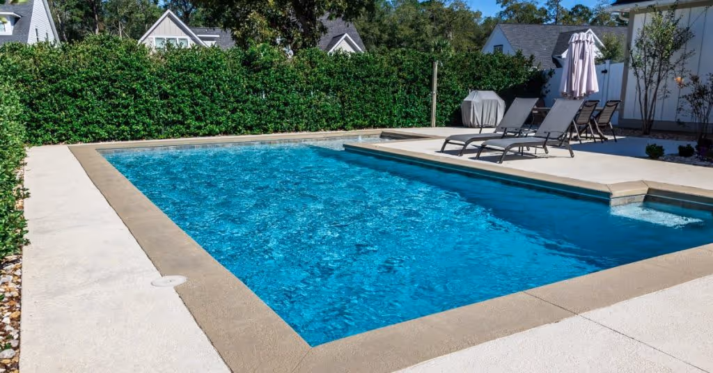 A swimming pool surrounded by green hedges. The pool area has lounge chairs and a gray, folded umbrella.