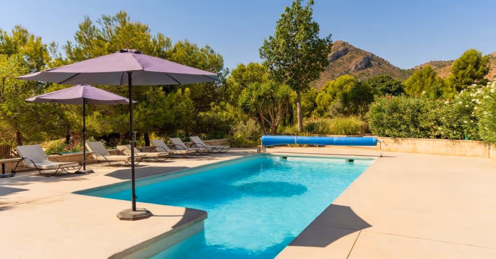 A swimming pool with purple umbrellas, a line of cushioned lounge chairs, and greenery and mountains in the background.