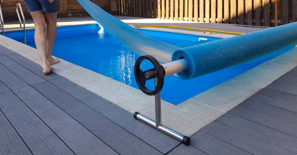 A person unravels a pool cover and pulls it over the pool. The swimming pool is enclosed with a brown fence.