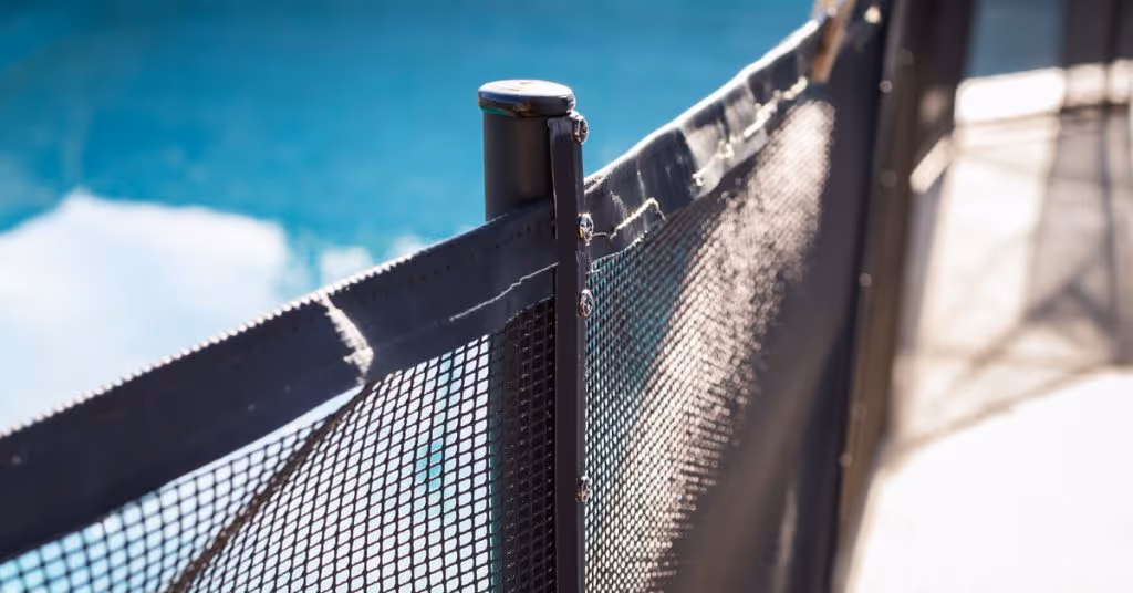 A black mesh swimming pool fence is around a backyard pool. It’s daytime and sunlight illuminates the pool fence.