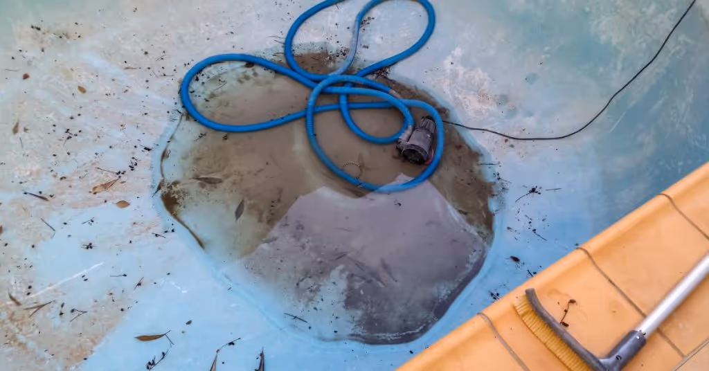 Bottom of a drained swimming pool with a blue curled vacuum pump. The surface has a small puddle and debris.
