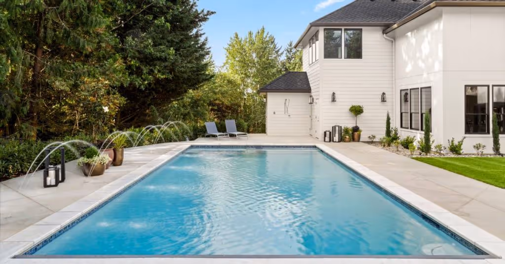 A close-up of a luxurious home exterior and a large swimming pool on a sunny day. The pool has water jets forming arches.