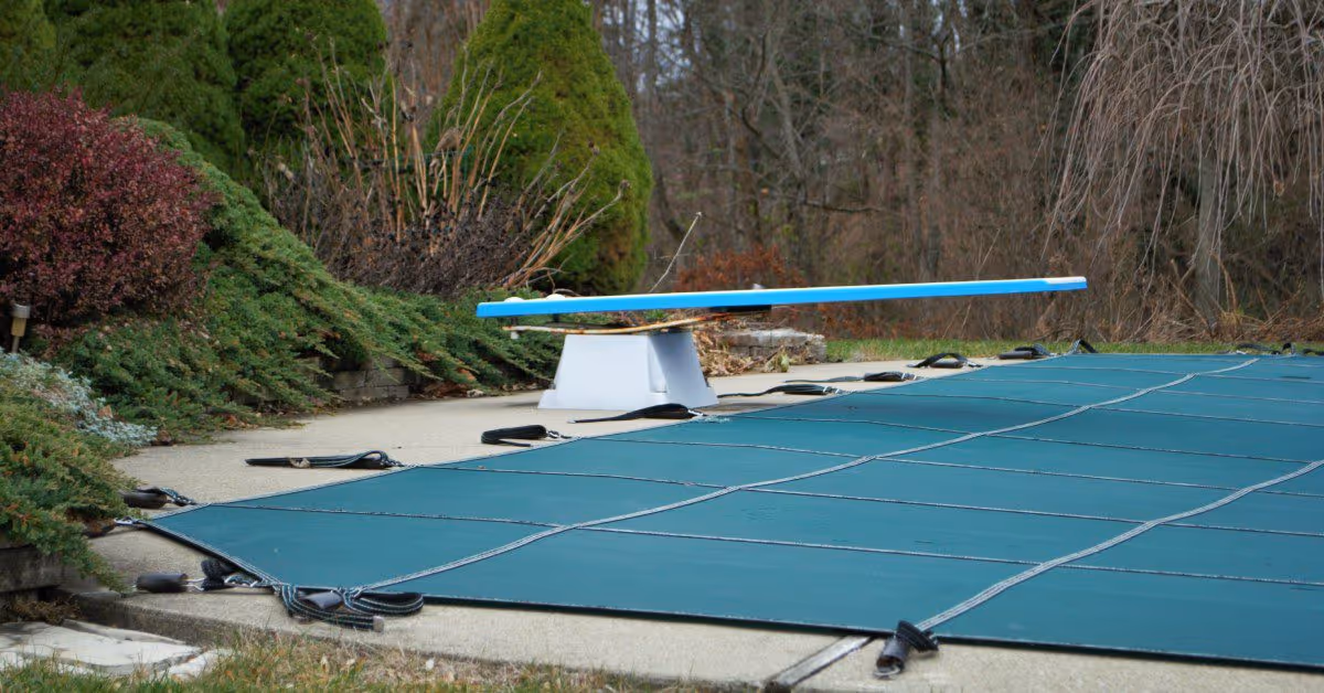 A backyard swimming pool with a green cover. Above the cover is a blue diving board. Greenery surrounds the area.