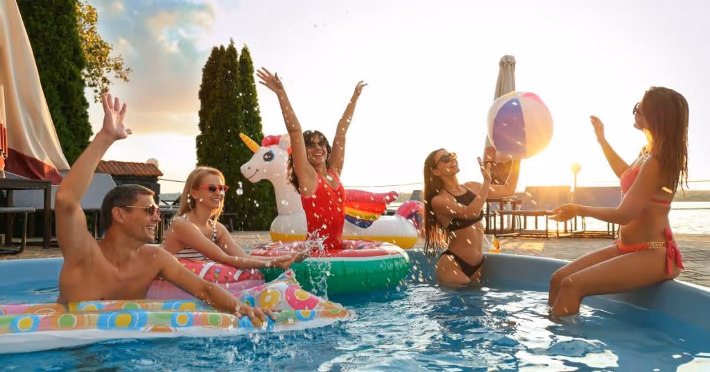 A group of happy people in a pool. One woman tosses a beach ball to another woman, and some people lounge on pool floaties.