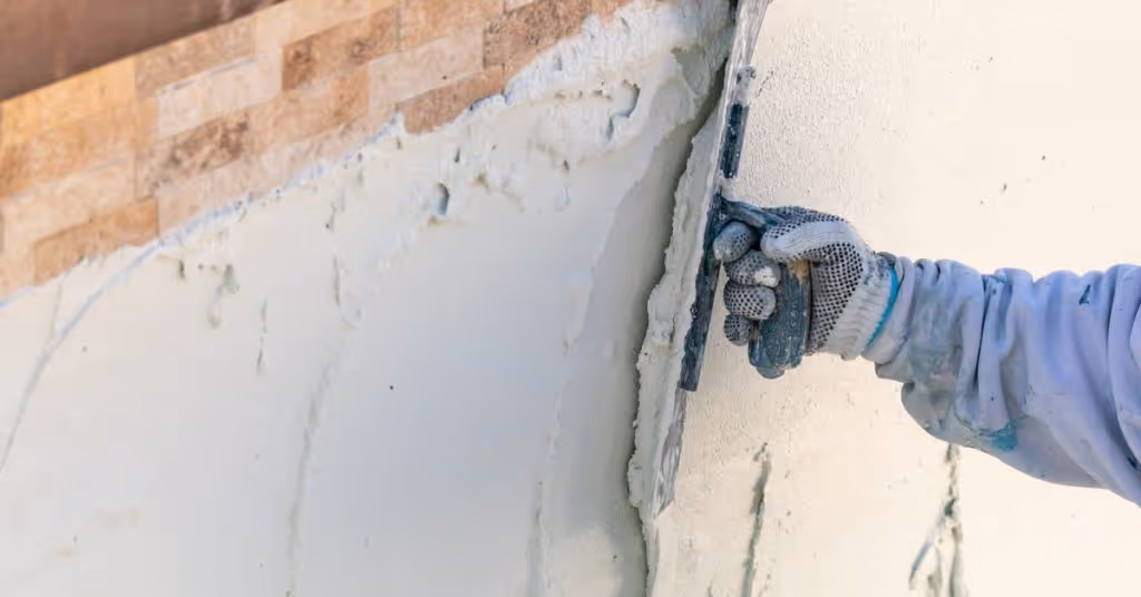 A close-up of someone's hand replastering a backyard pool with a trowel. The pool has beige and brown tile.