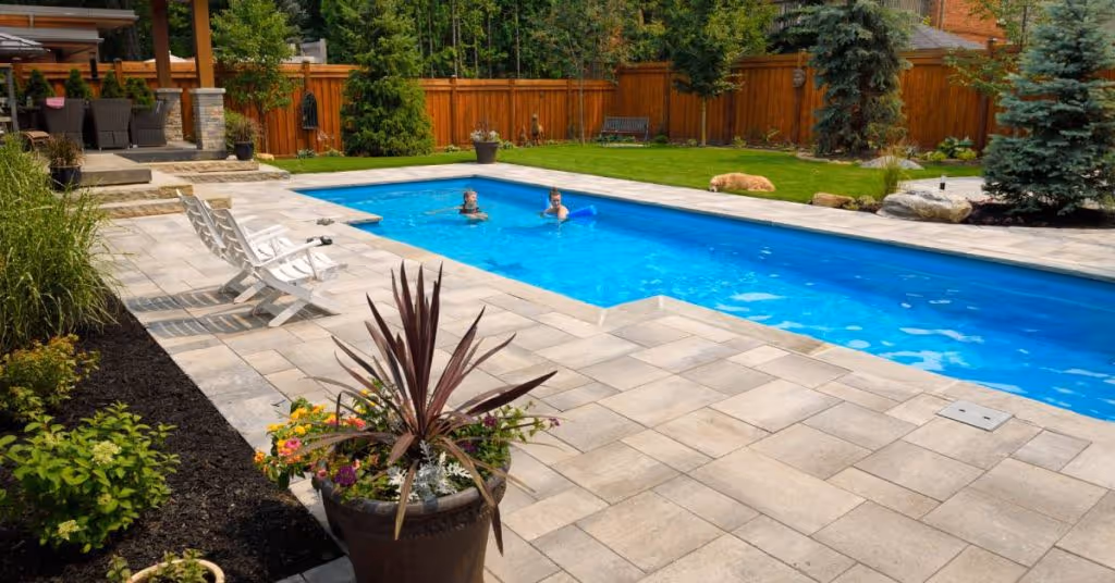 Two people are swimming in a brand-new backyard pool with their dog. Patio pavers and a green lawn surround the pool.