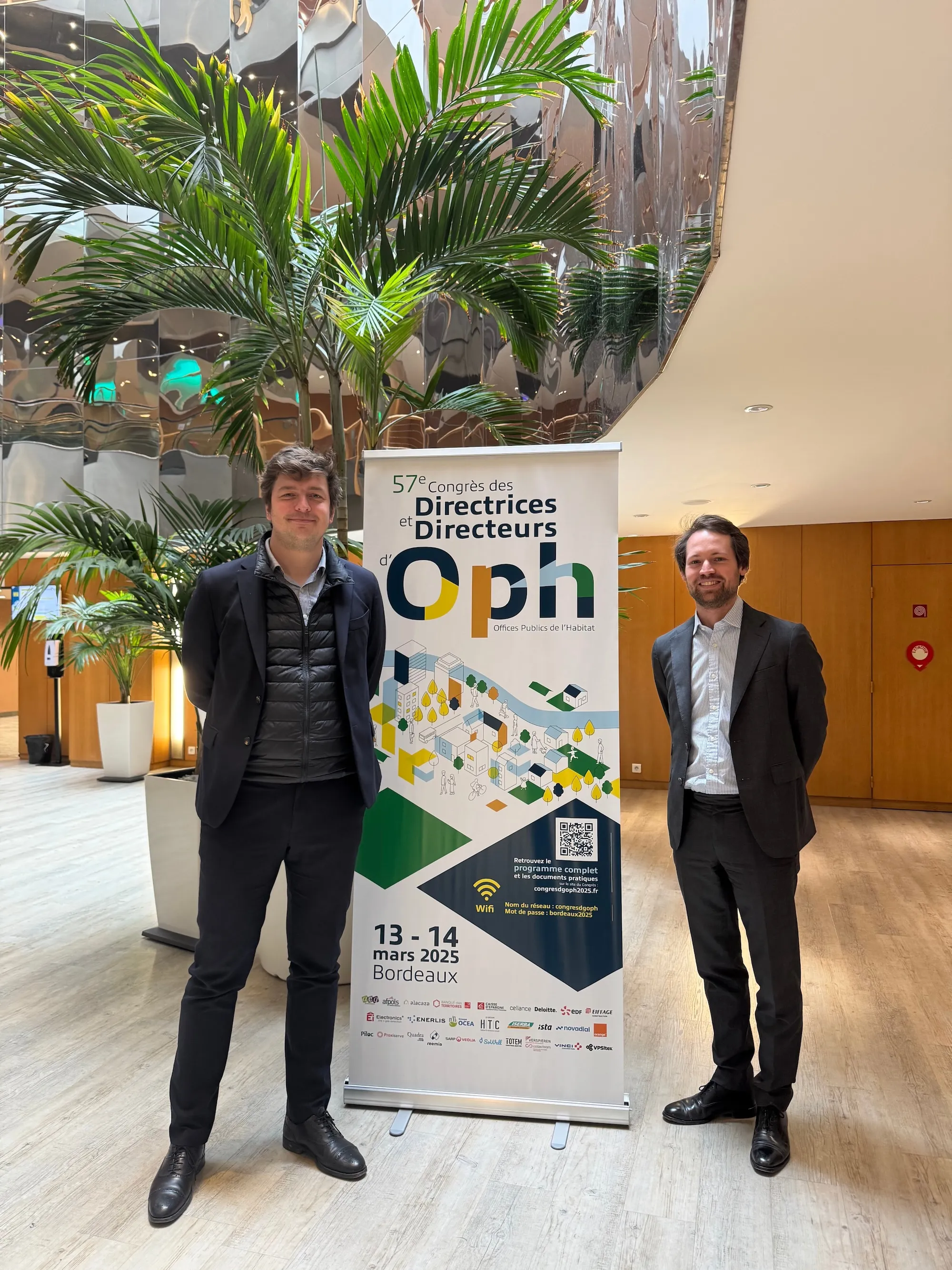 Deux hommes en costume posent devant un kakémono du congrès des Directrices et Directeurs d’OPH, illustrant la participation de Piloc à l’événement.