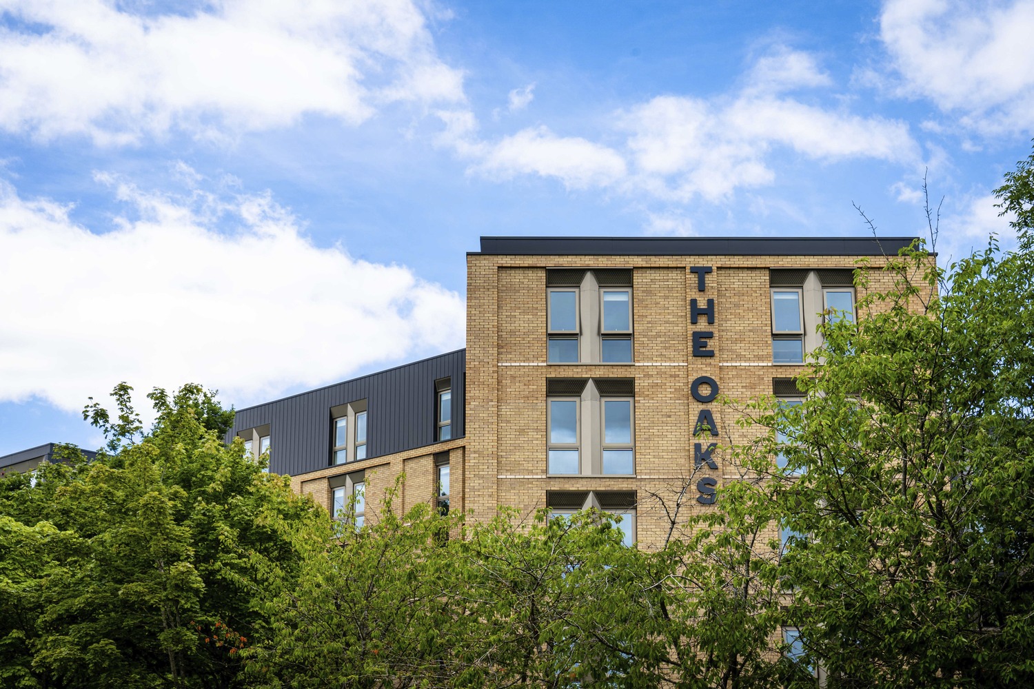 The Oaks student accommodation, a buff brick building with its name on the facade, behind trees