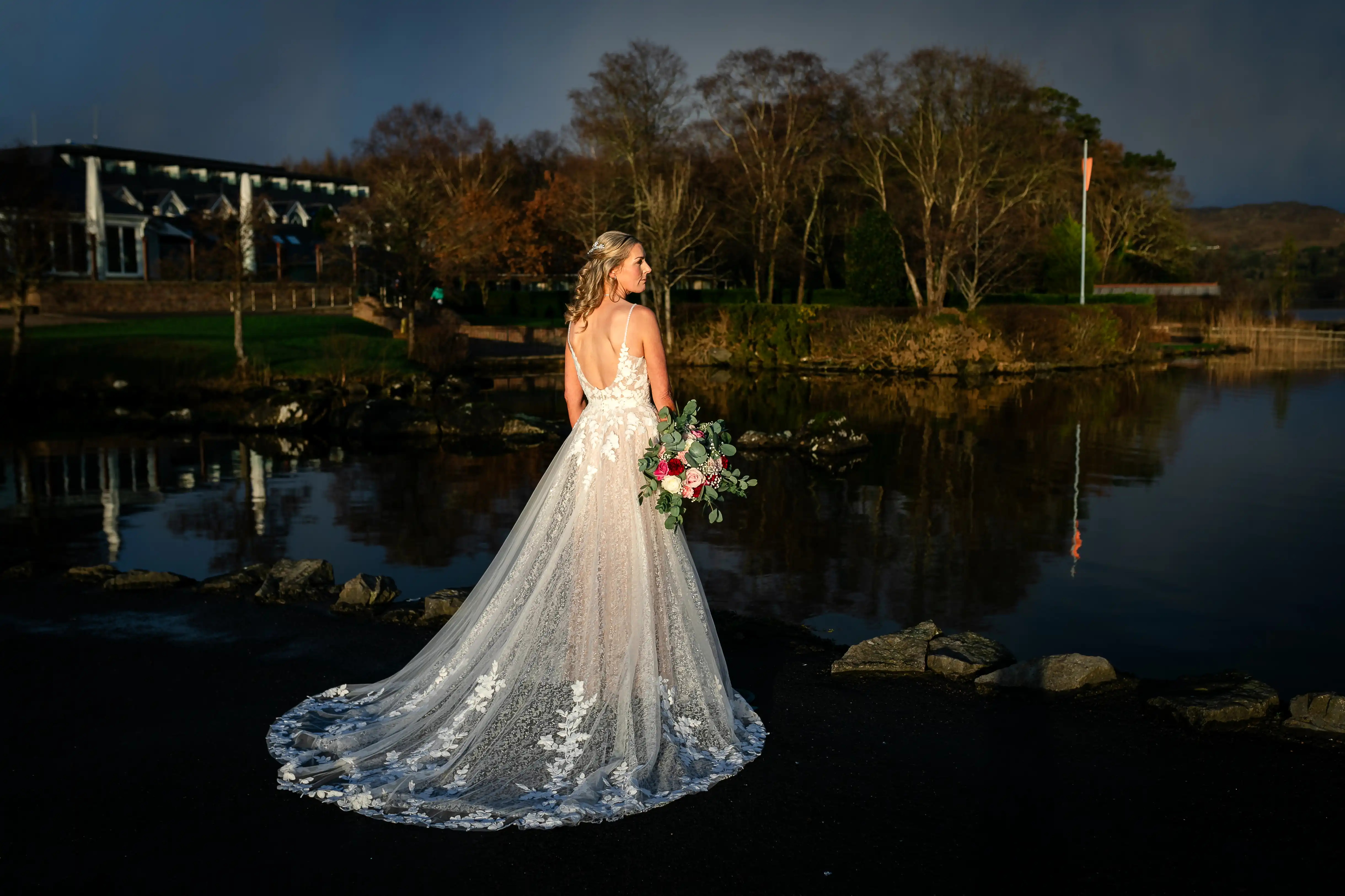 Bride in dress holding flowers in front of a lake facing away from camera