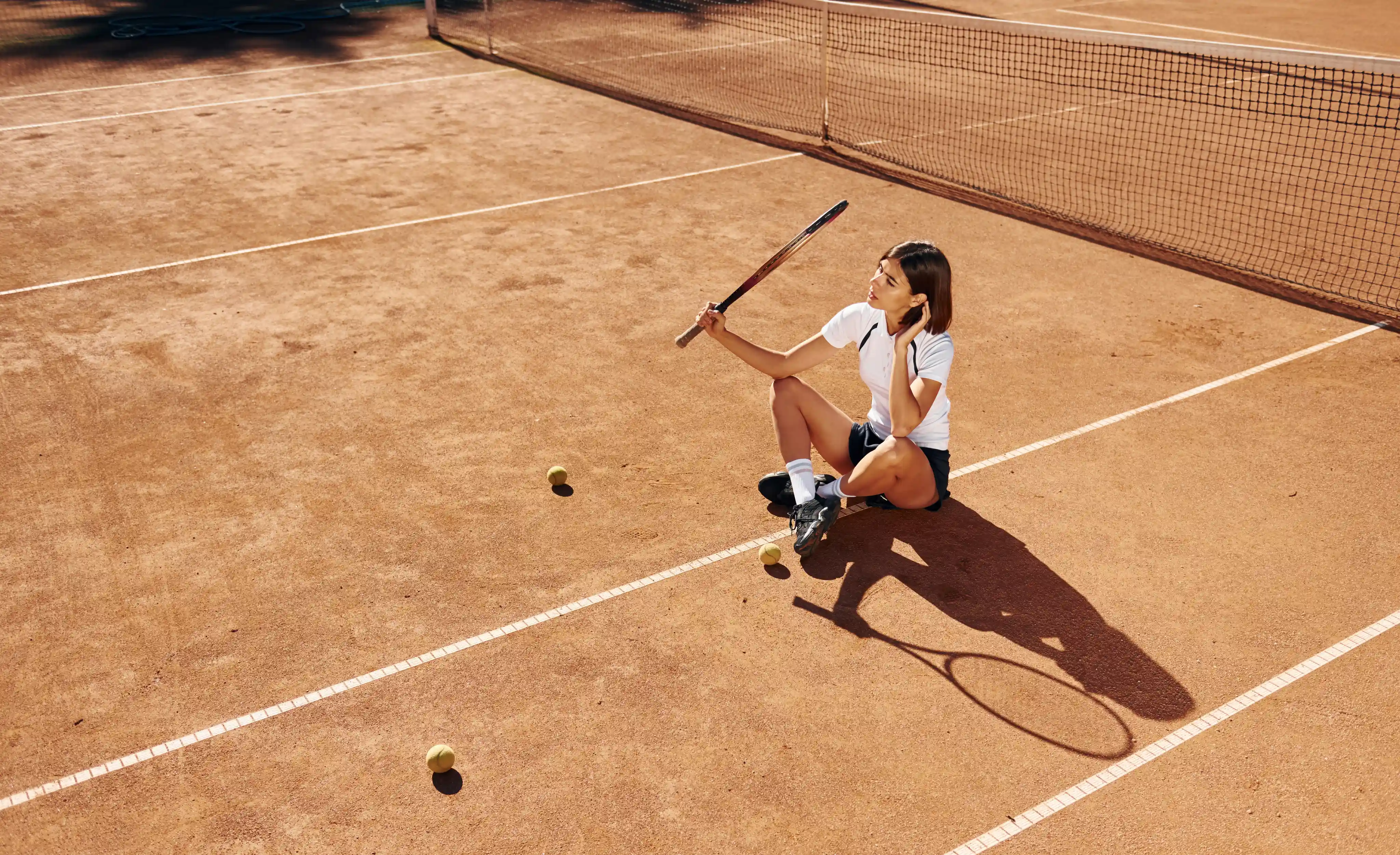 A tennis player sits on the clay court, holding a racket and a phone, surrounded by scattered tennis balls in bright sunlight.