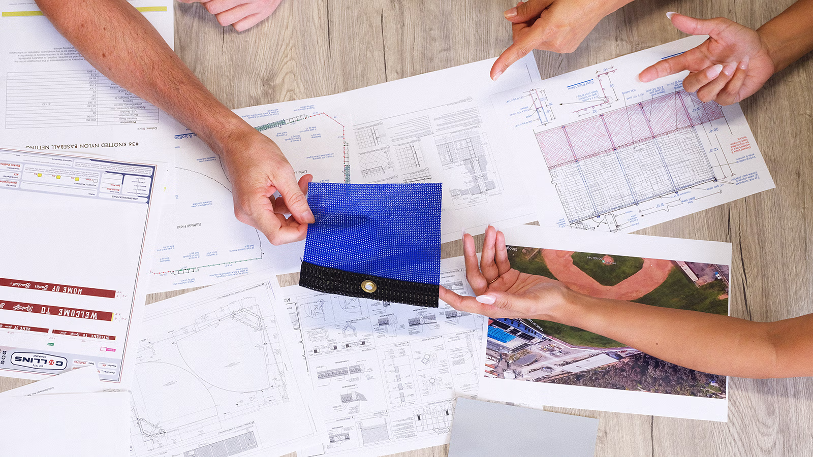 Three professionals discuss blueprints on a table, with a safety helmet and a tablet nearby, as one gestures while speaking.