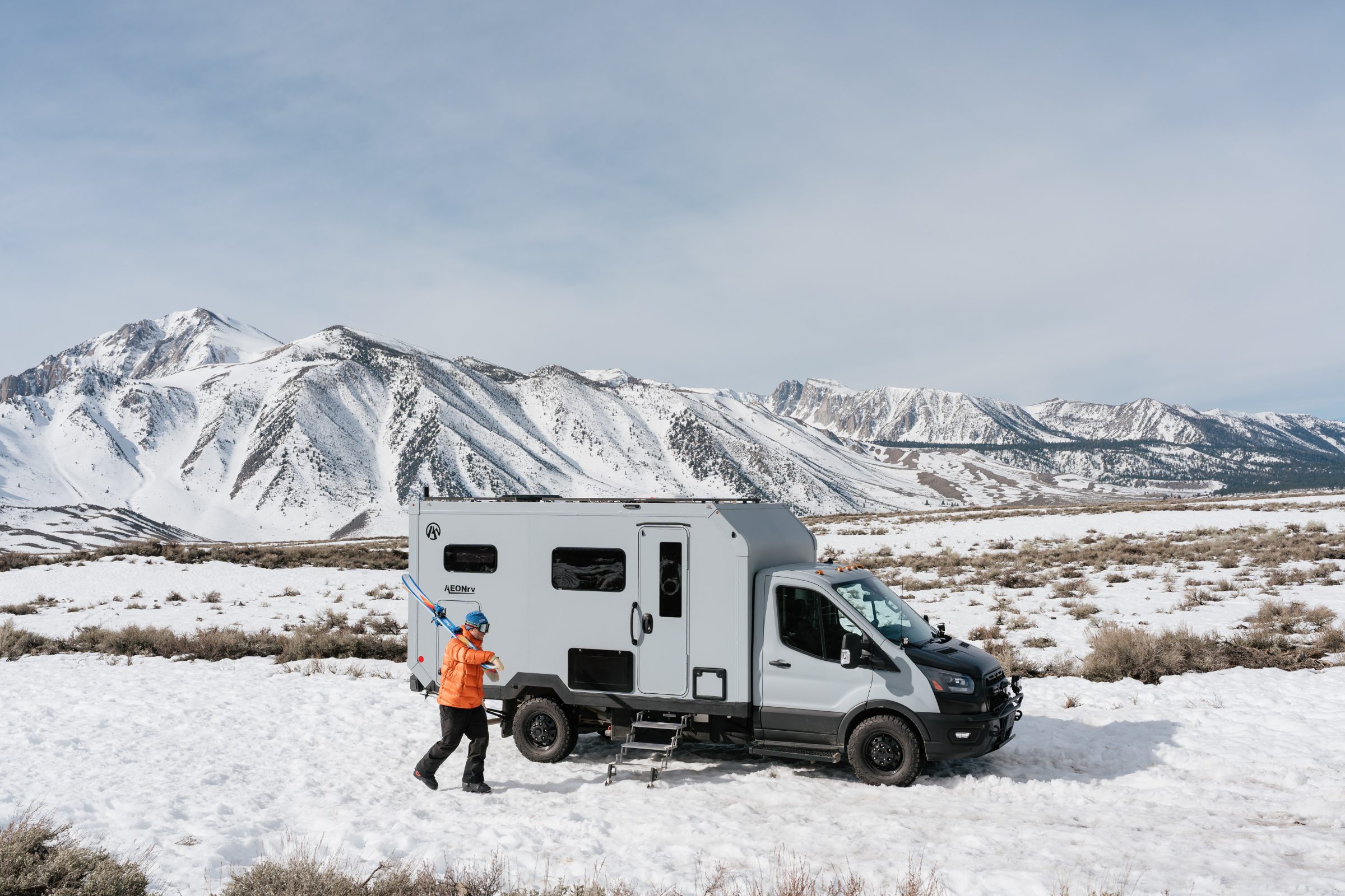 Skier carrying skis to the AEONrv parked in snow below mountain peaks