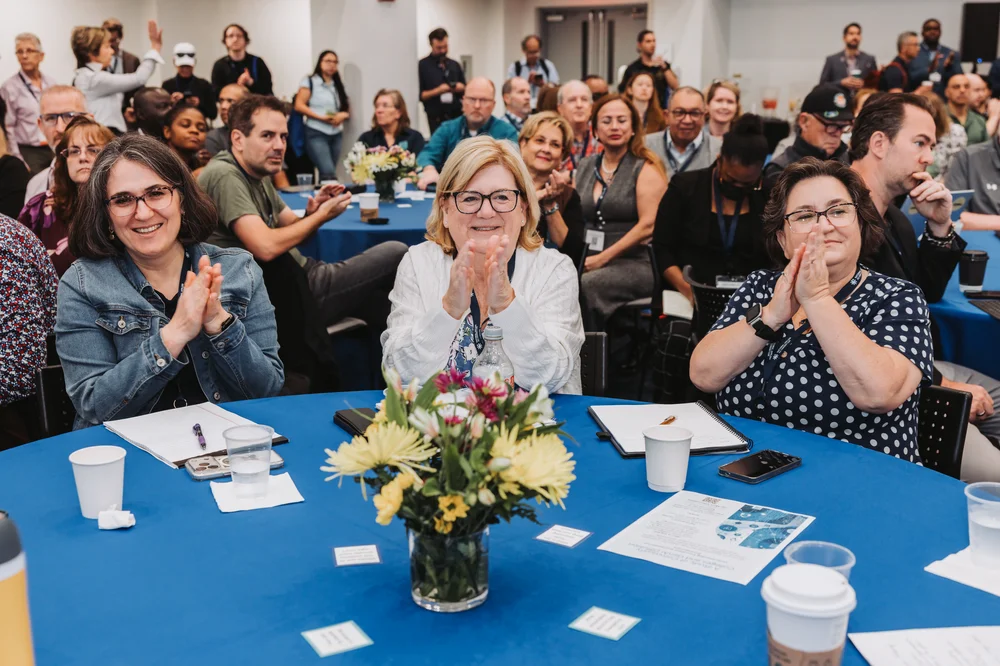 Audience of diverse adults sitting at round tables with blue tablecloths, clapping and smiling during an indoor event.