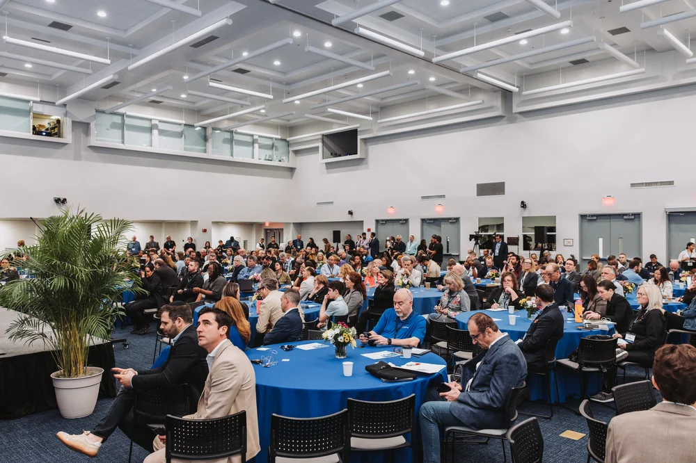 Large group of people seated at round tables with blue tablecloths in a conference room attending an event.