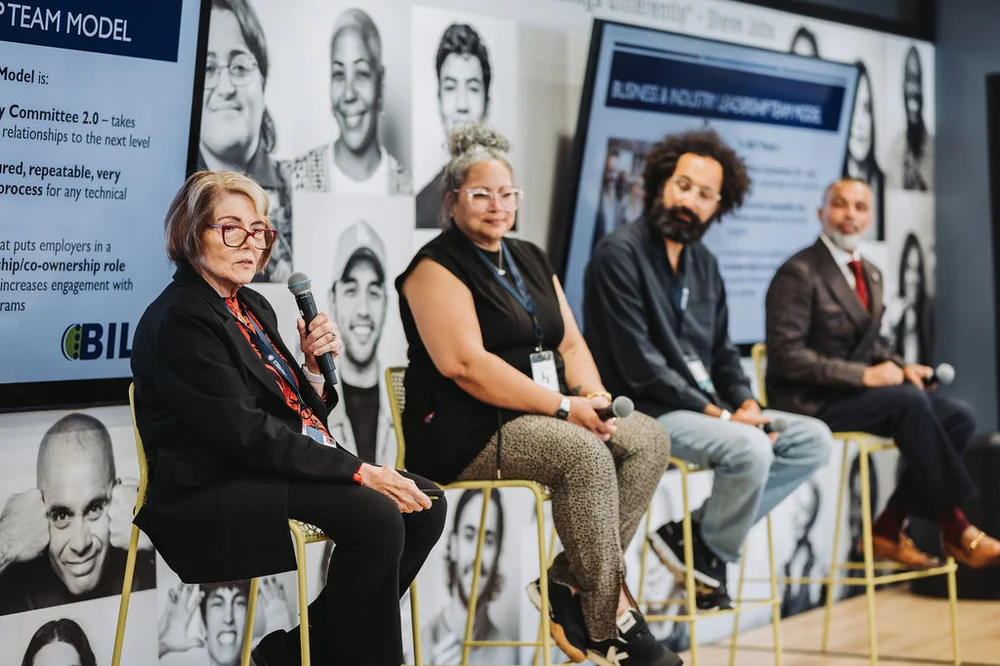 Four people sitting on high chairs in a panel discussion with a screen and black-and-white portraits behind them.