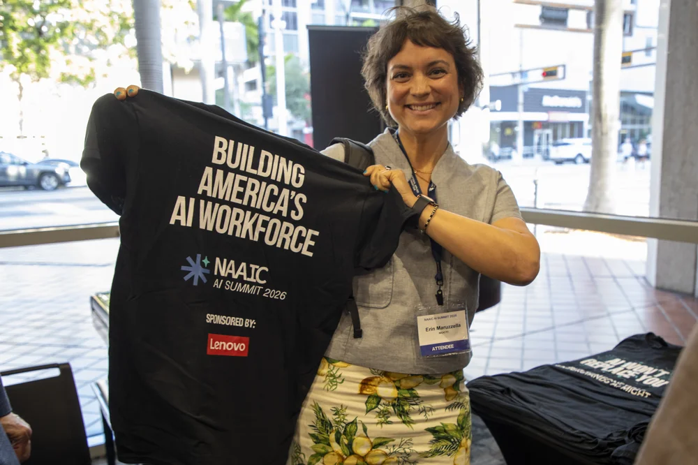 Smiling woman holding up a black t-shirt with the text 'Building America's AI Workforce, NAAIC AI Summit 2026, Sponsored by Lenovo.'