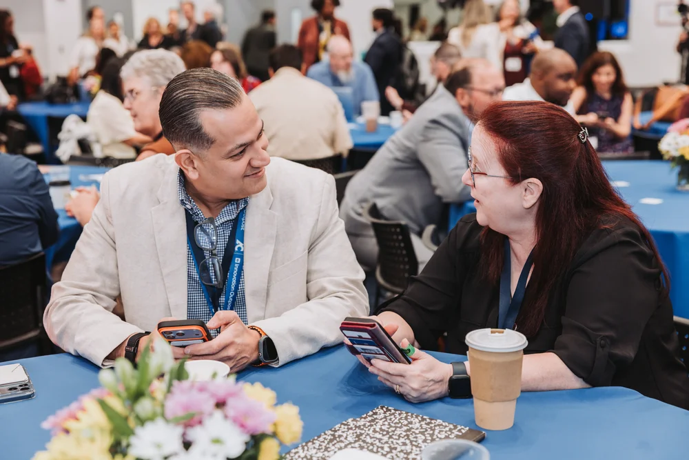 Two people sitting at a table covered with a blue cloth, smiling and holding phones, with a coffee cup and flowers in the foreground and others socializing in the background.