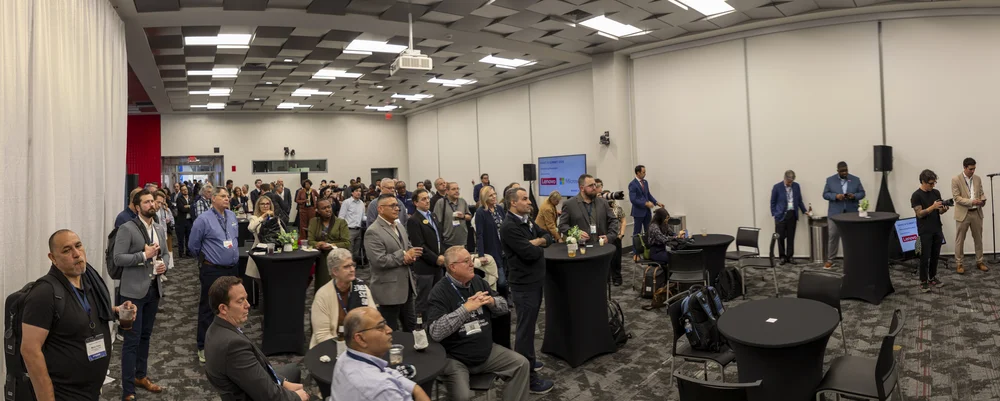 Group of people attentively listening during a conference or presentation in a large room with tables and chairs.