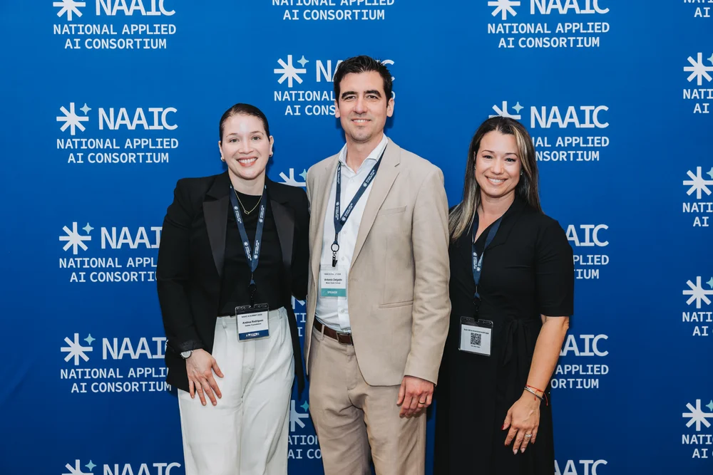 Three people smiling and standing in front of a blue National Applied AI Consortium (NAAIC) backdrop.