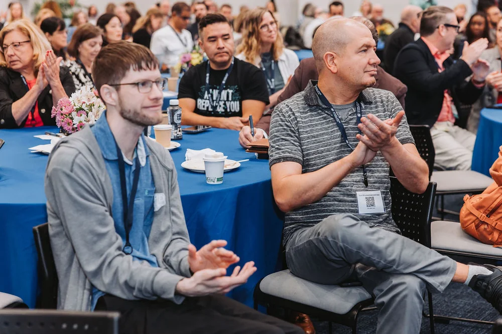 People sitting and clapping at a conference or seminar with blue tablecloths and name badges.