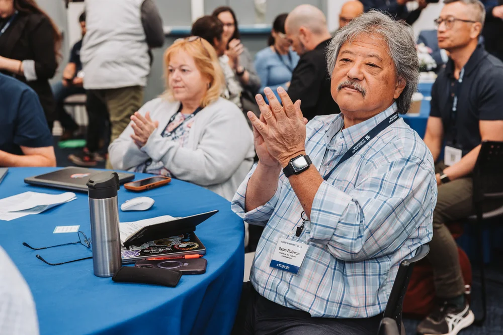 An older man with gray hair and a mustache clapping while seated at a conference table with a blue tablecloth, surrounded by other attendees.
