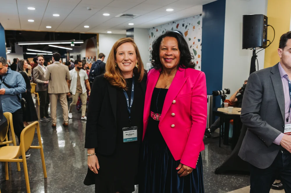 Two women smiling and posing for a photo at a professional event with people and chairs in the background.