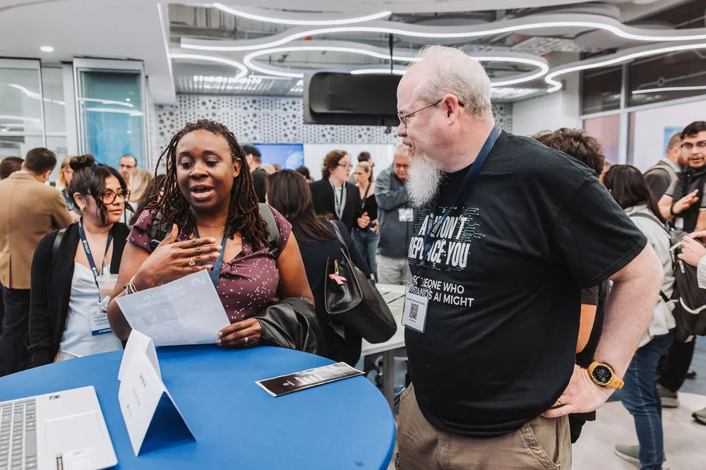 A woman and a man engaged in conversation at a blue round table with a laptop in a busy conference room.