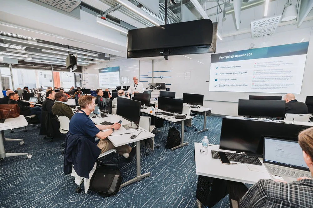 Modern classroom with students seated at desks using computers and a presenter standing near a large screen displaying 'Prompting Engineer 101'.