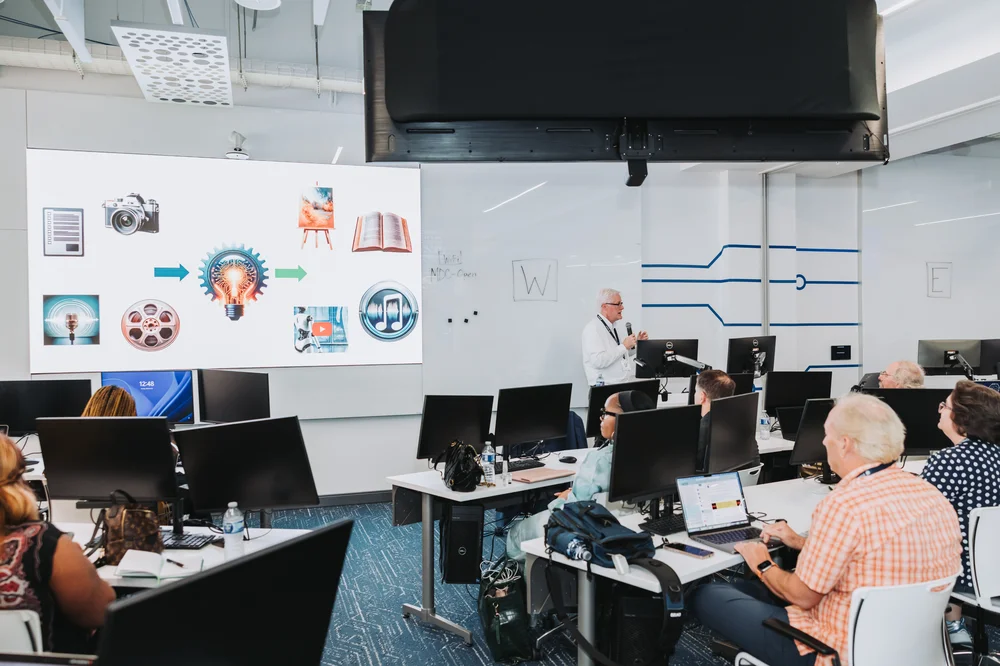 Instructor presenting a digital media concept with icons on a large screen to an attentive adult classroom.