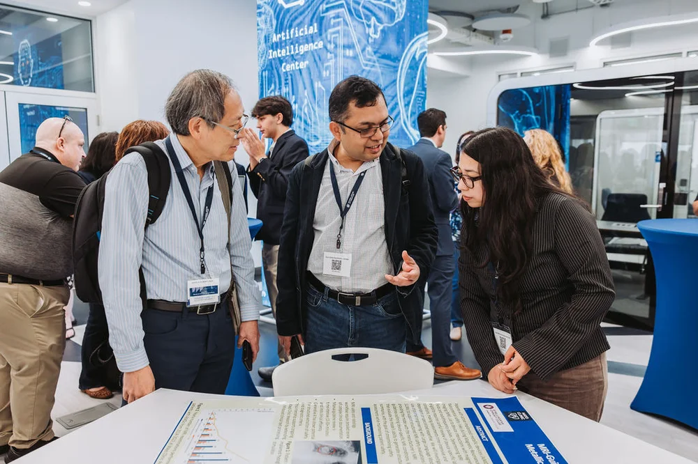 Three people discussing a research poster at an Artificial Intelligence Center event in a modern indoor setting.