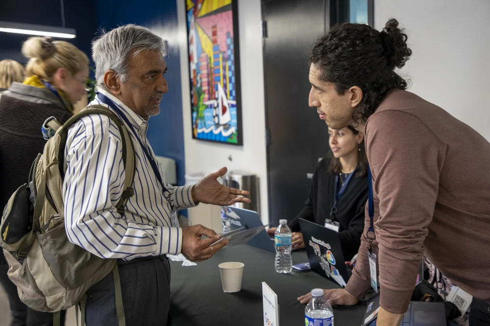 Older man with a backpack talking to a young man leaning on a table at an event booth with laptops and bottled water.