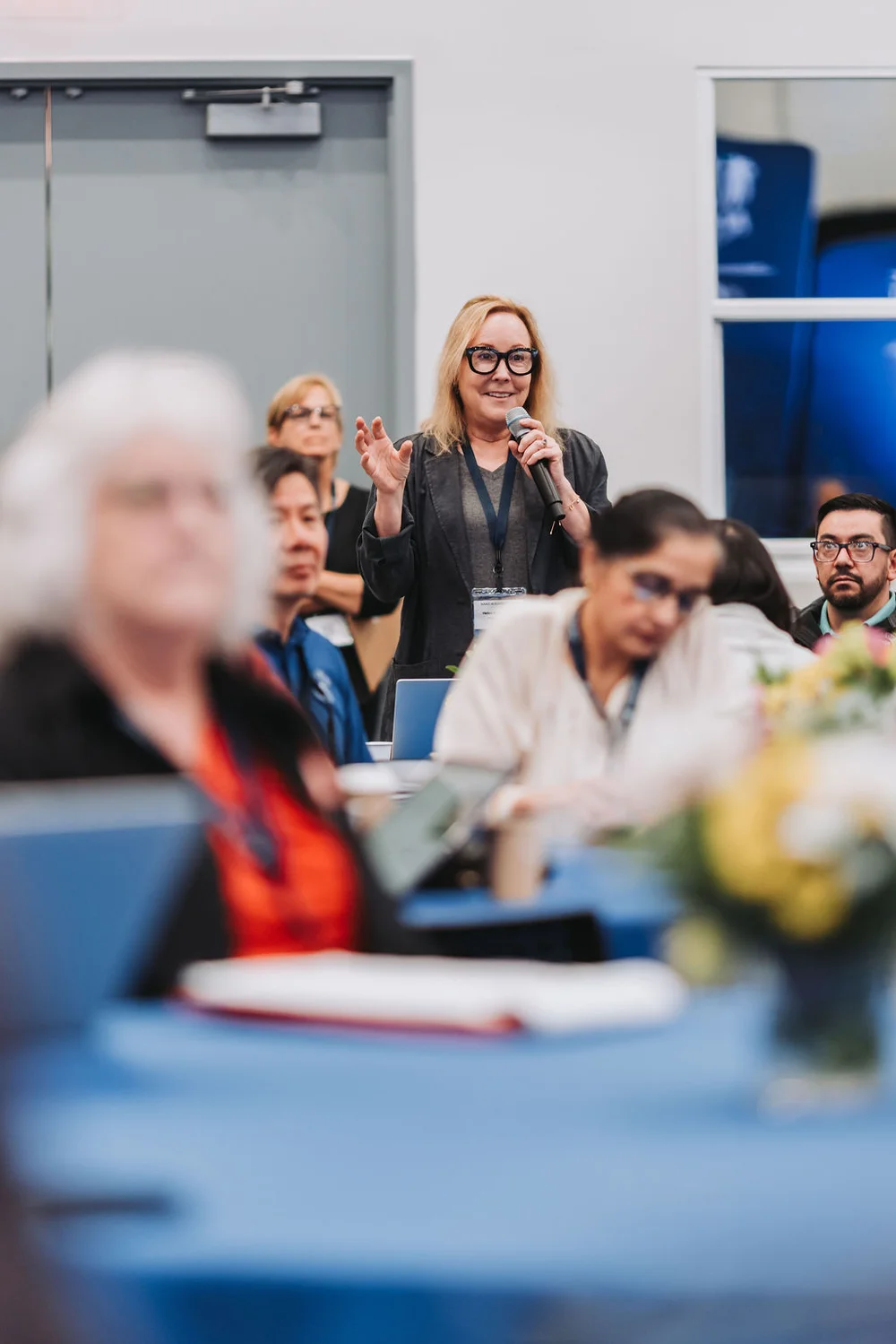 Woman in glasses speaking into a microphone at a conference with attendees listening in the background.