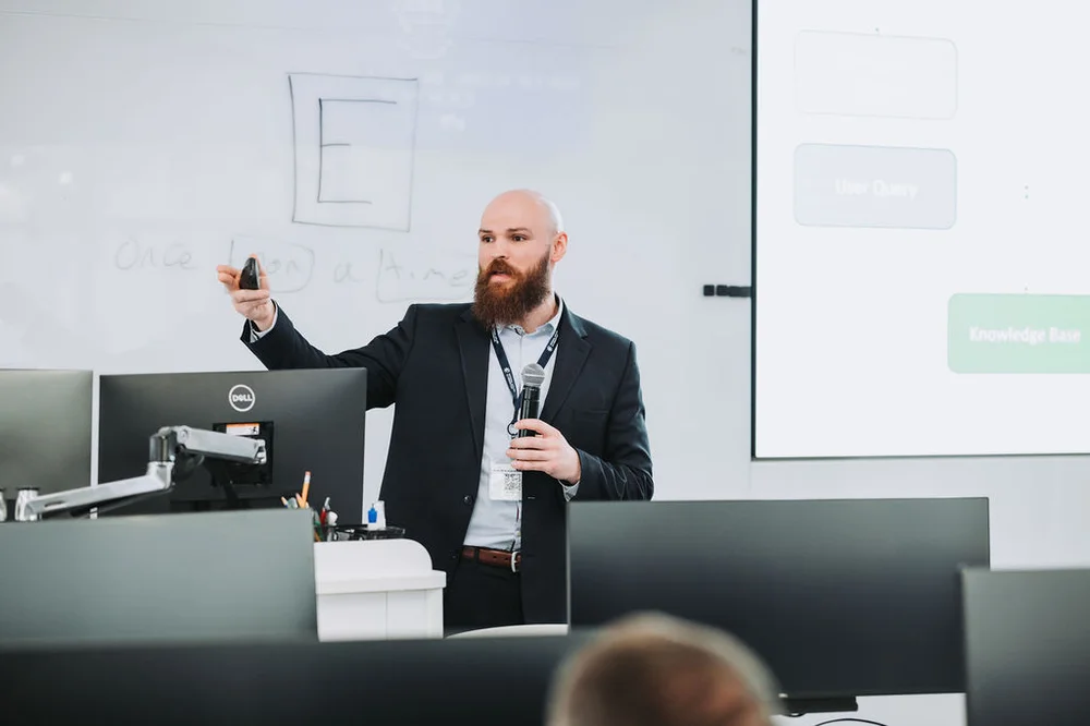 Bearded man in a suit holding a microphone and pointing at a whiteboard during a presentation in a conference room.