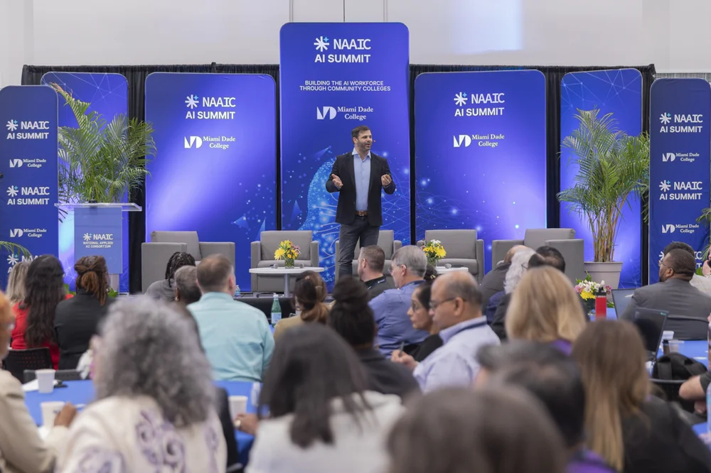 Speaker presenting on stage at NAALC AI Summit hosted by Miami Dade College with an audience seated in front.