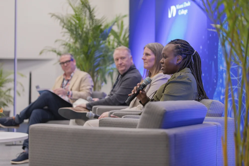 Panel of four people sitting on stage armchairs, one woman speaking into a microphone during a discussion event.