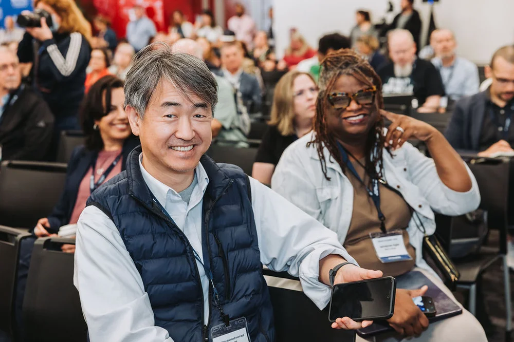 Smiling man showing a smartphone screen while sitting next to a woman wearing sunglasses at a conference audience.