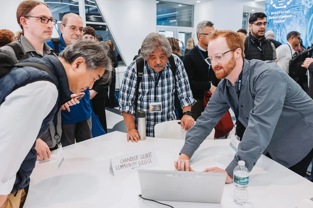 Group of people gathered around a table with a laptop, discussing in a conference setting with a sign reading Chandler Gilbert Community College.