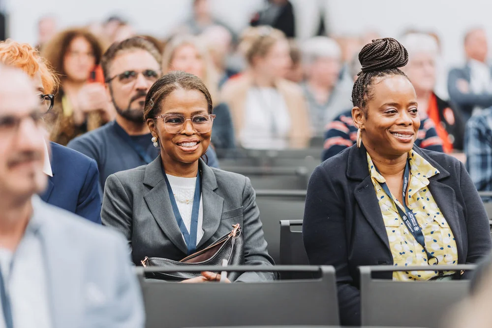 Two professional women sitting and smiling in an audience at a conference or seminar.