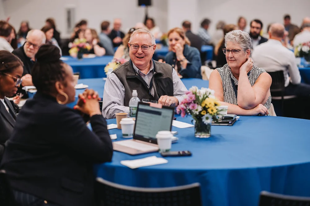 Group of people sitting at a round table with blue tablecloth, engaged in conversation during an indoor event.