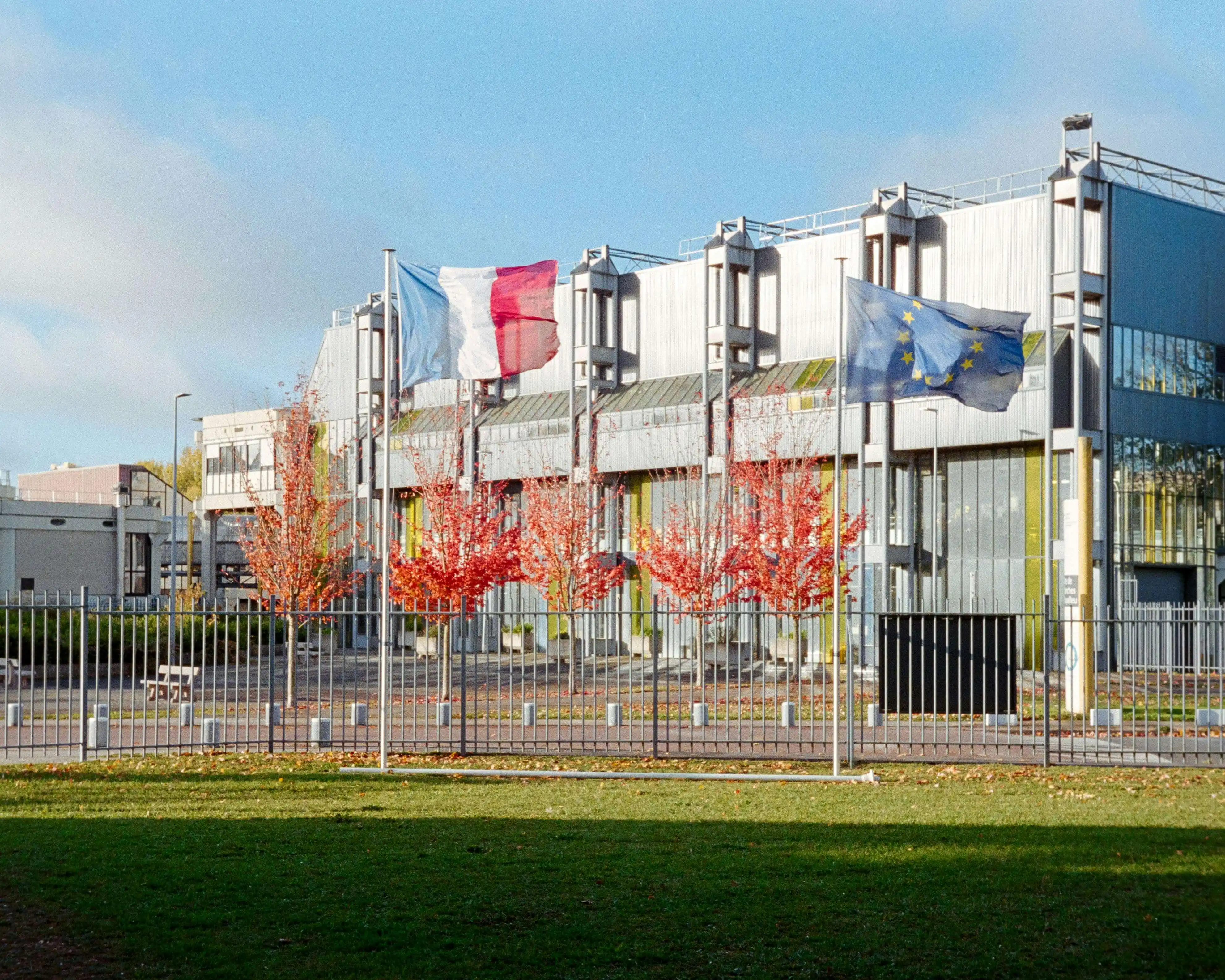 Bâtiment industriel moderne avec drapeaux français et européen flottant devant des arbres rouges en automne.