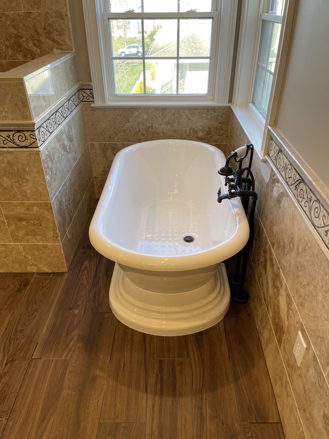 Freestanding pedestal soaking tub in a Stewartsville, NJ bathroom on a wood-look plank tile floor, with an oil-rubbed bronze floor-mounted filler and a mosaic tile border on the wall behind