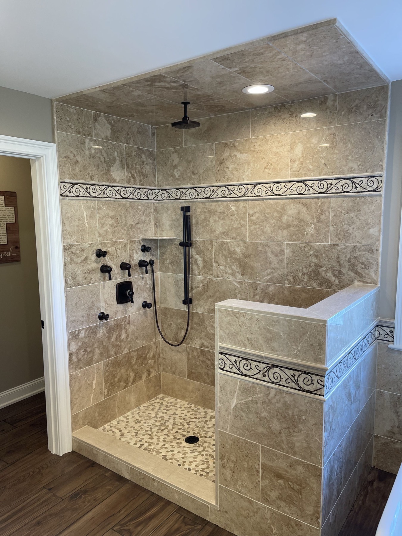 Walk-in shower in a Stewartsville, NJ primary bathroom with travertine-look porcelain walls, a scroll mosaic border, pebble tile floor and oil-rubbed bronze rain head and body sprays