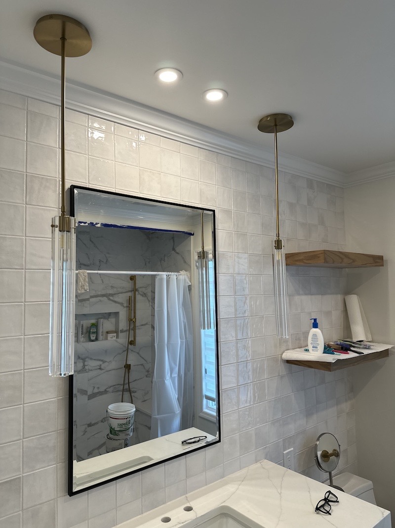 Bathroom vanity wall in Pittstown, NJ with square stacked white tile, brass and glass pendant lights, floating wood shelves and a black-framed mirror